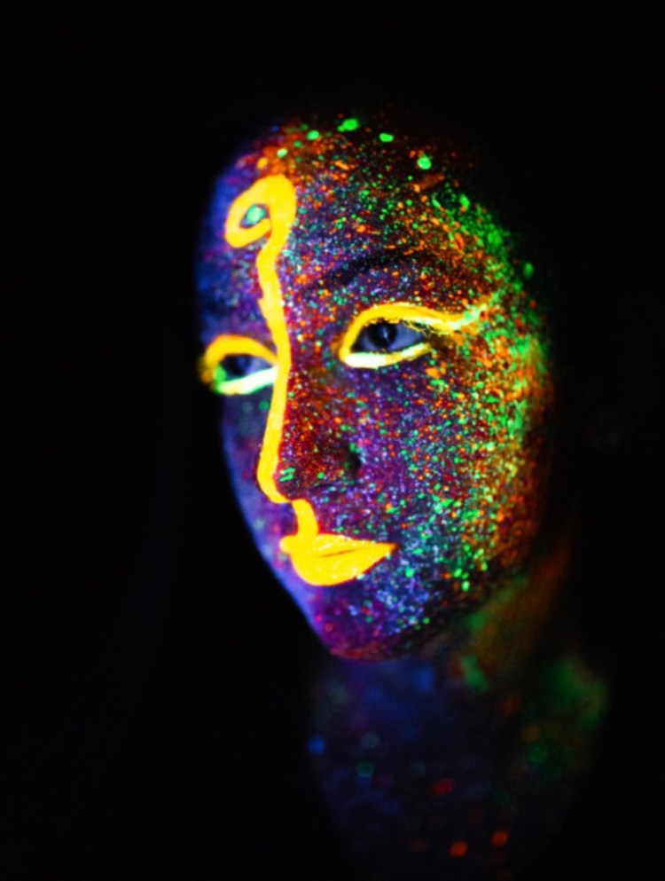 The image shows face of a woman. It is painted with luminous glitter paint and the features are highlighted with bright yellow lines, ending in a question mark on the forehead.